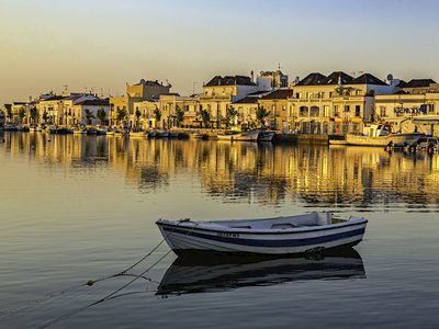Small boat docked with houses in background reflecting golden glow onto calm waters, Tavira, Portugal