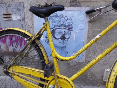 Rusty yellow bicycle leaning against wall in San Sepolcro which has a street art painting rendition on wall of David of Michelangelo statue depicting David underwater wearing scuba mask, Italy