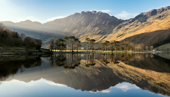 Calm waters of Buttermere Lake reflecting Haystacks fell towering from behind with sun shining through blue skies, Lake District, England