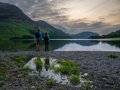 Male and female walkers at edge of Buttermere Lake taking in view of Lake District at sunset