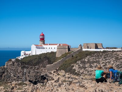 Two hikers sat on rocks taking in distant view of Sagres lighthouse on sunny day, Algarve, Portugal