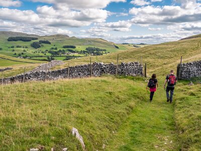 Two walkers enjoying hike in settle countryside walking towards opened gate amidst grassy hills, Yorkshire Dales