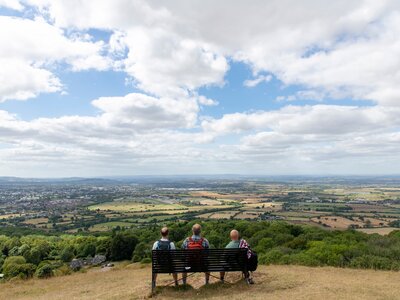 Three hikers sitting on bench on top of Cleeve Hill along the public footpath Cotswold way with panoramic view on the valley and outskirts of city of Cheltenham, UK 